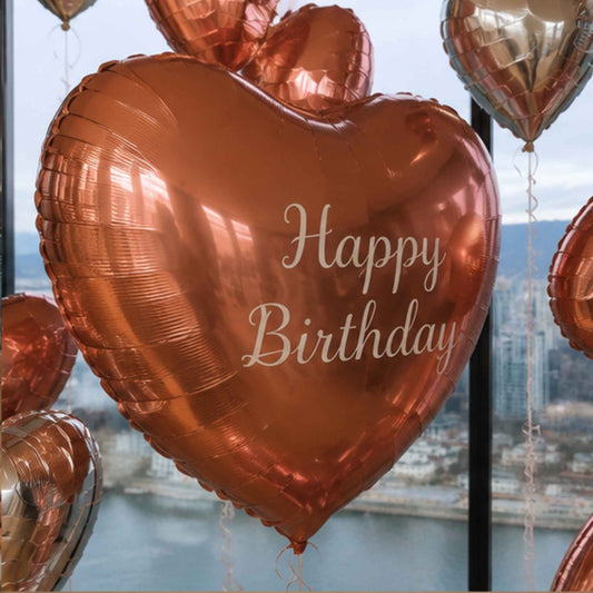 A metallic rose gold heart-shaped foil balloon with 'Happy Birthday' printed in white script, floating in front of a window with a city skyline and water in the background.