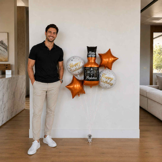 Man standing next to a decorative arrangement of balloons and a bottle with 'Happy Birthday' text in a modern indoor setting.