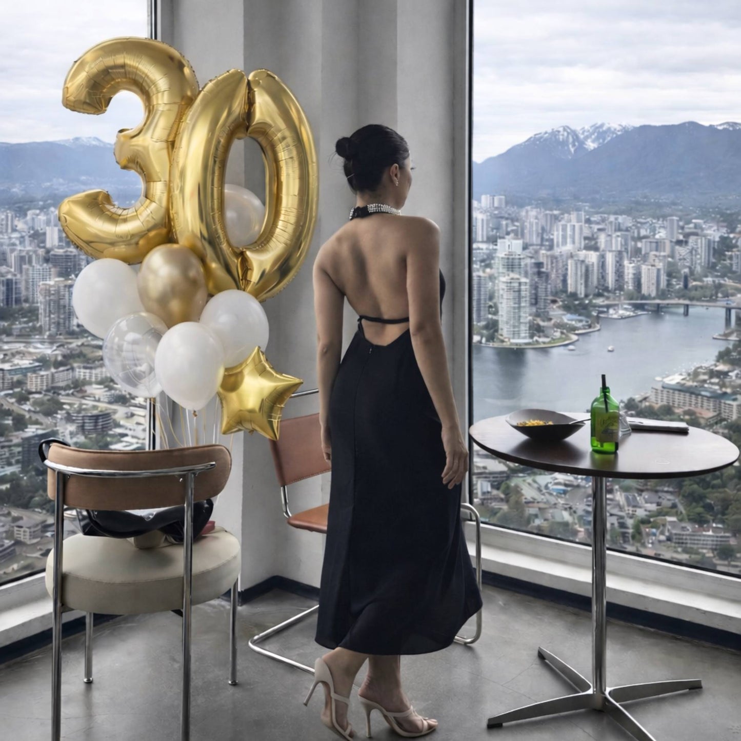 Woman in a black dress standing in a room with a cityscape view, next to gold '30' balloons.