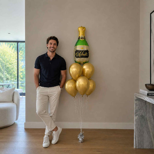 Man standing next to a large balloon shaped like a champagne bottle and bubbles in a living room.