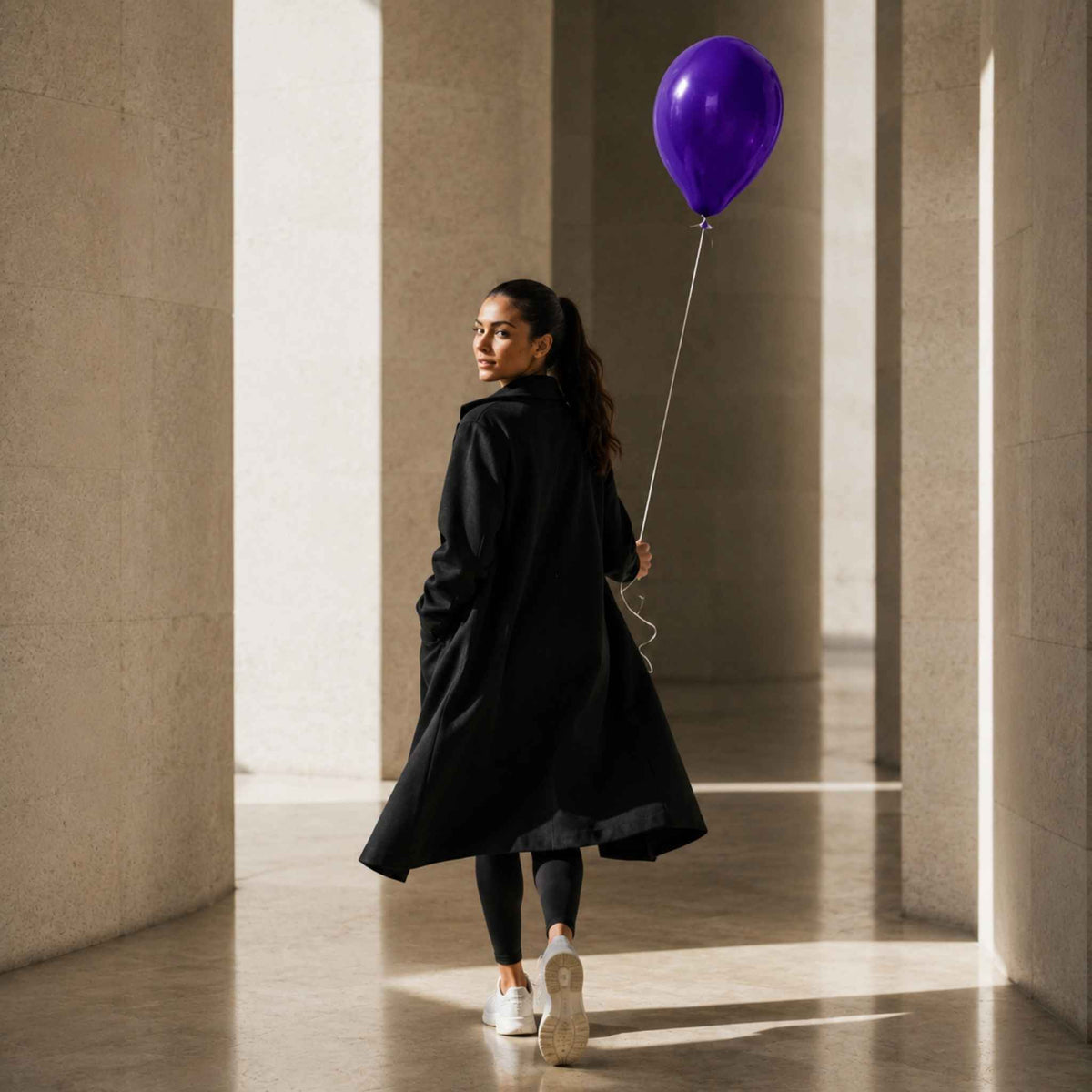 A woman in a long black coat, black leggings, and white sneakers walks through a sunlit stone corridor while holding a purple balloon.