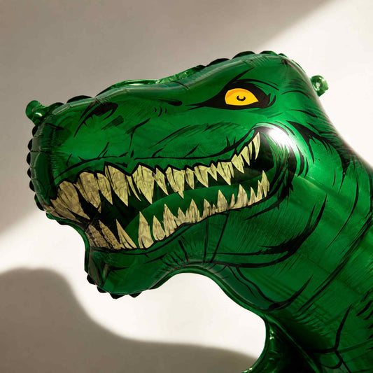 A green dinosaur head foil balloon with sharp white teeth and a yellow eye, photographed against a plain light background.