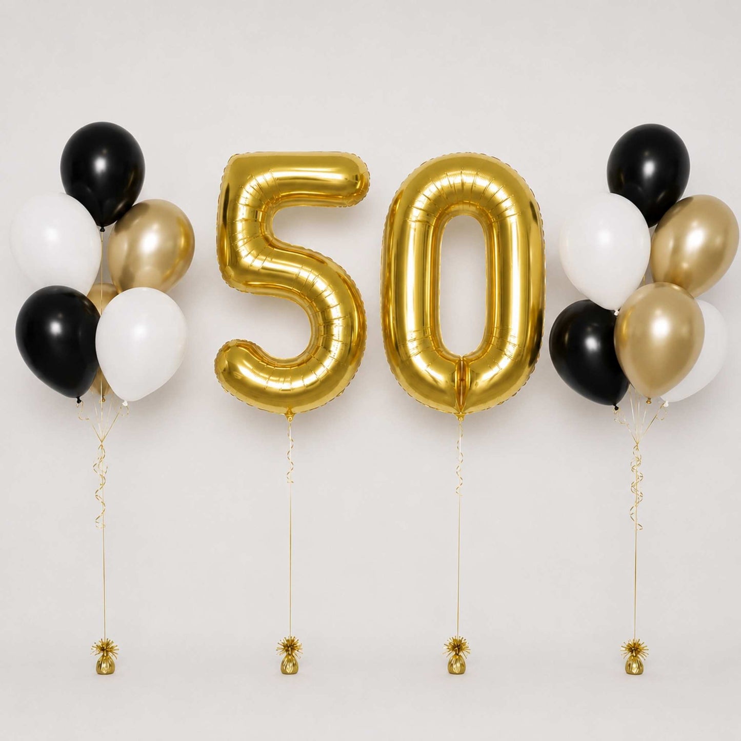 Gold foil number 50 balloons flanked by black, white, and gold balloon clusters, with small gold pineapple ornaments hanging from the strings against a plain light background.