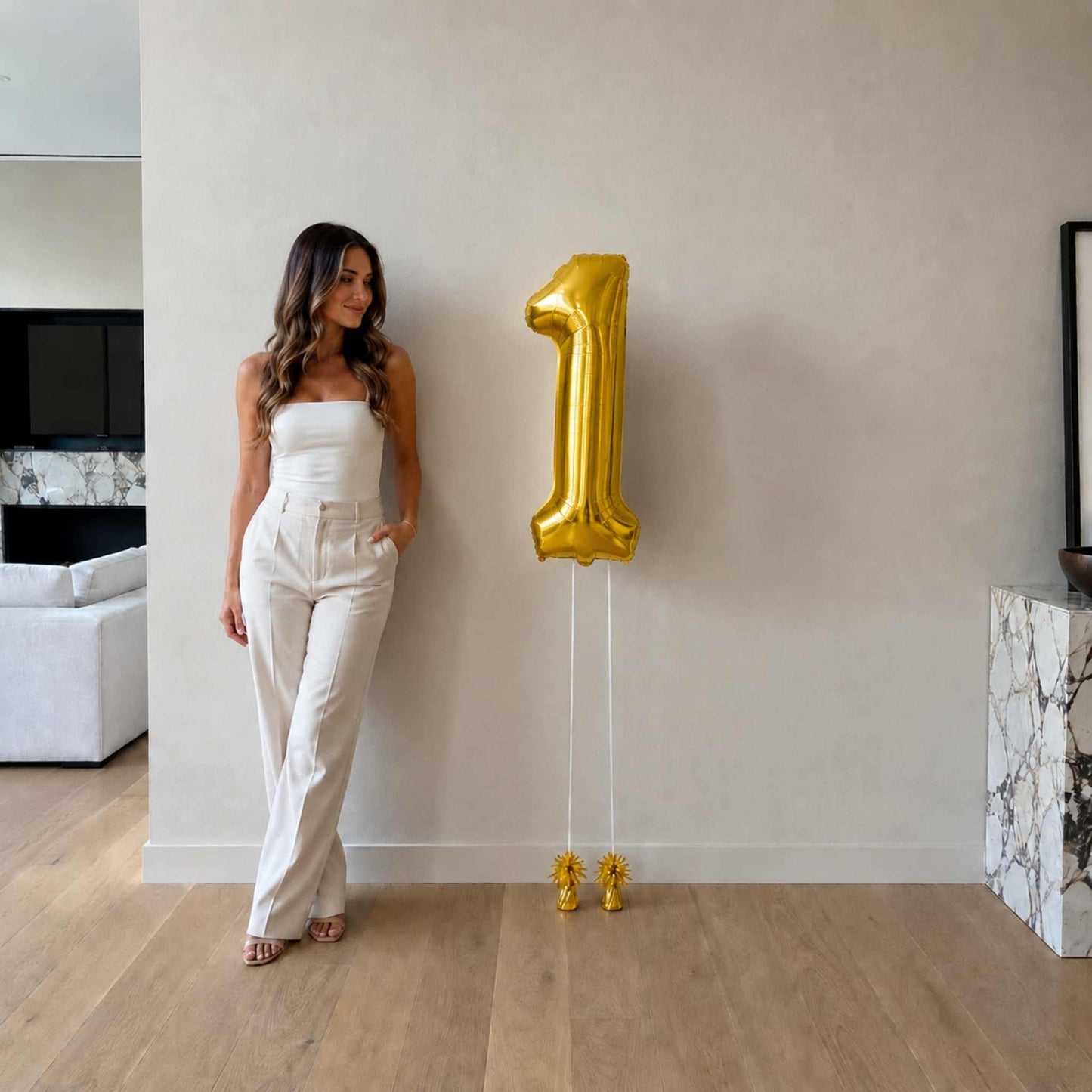 Woman in a white outfit standing next to a large gold number '1' balloon indoors.
