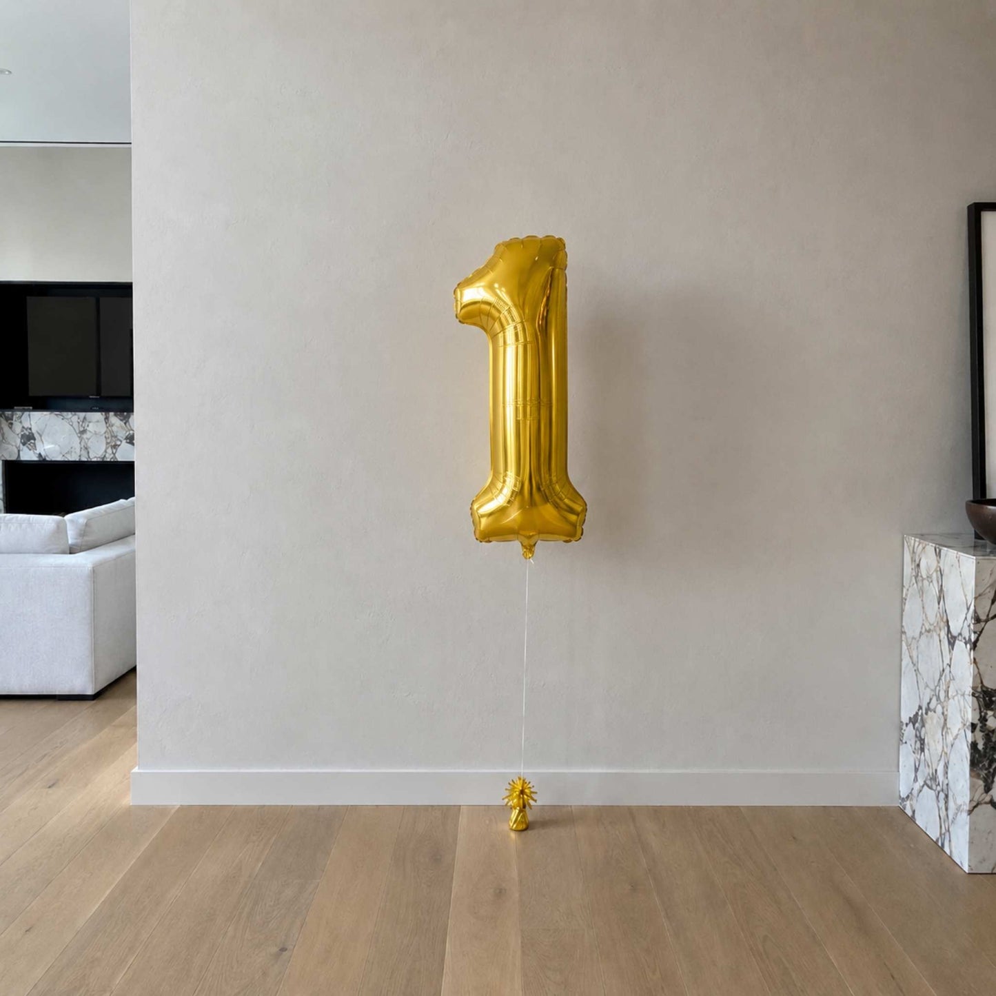 Gold number '1' balloon against a plain wall in a room with a couch and fireplace.