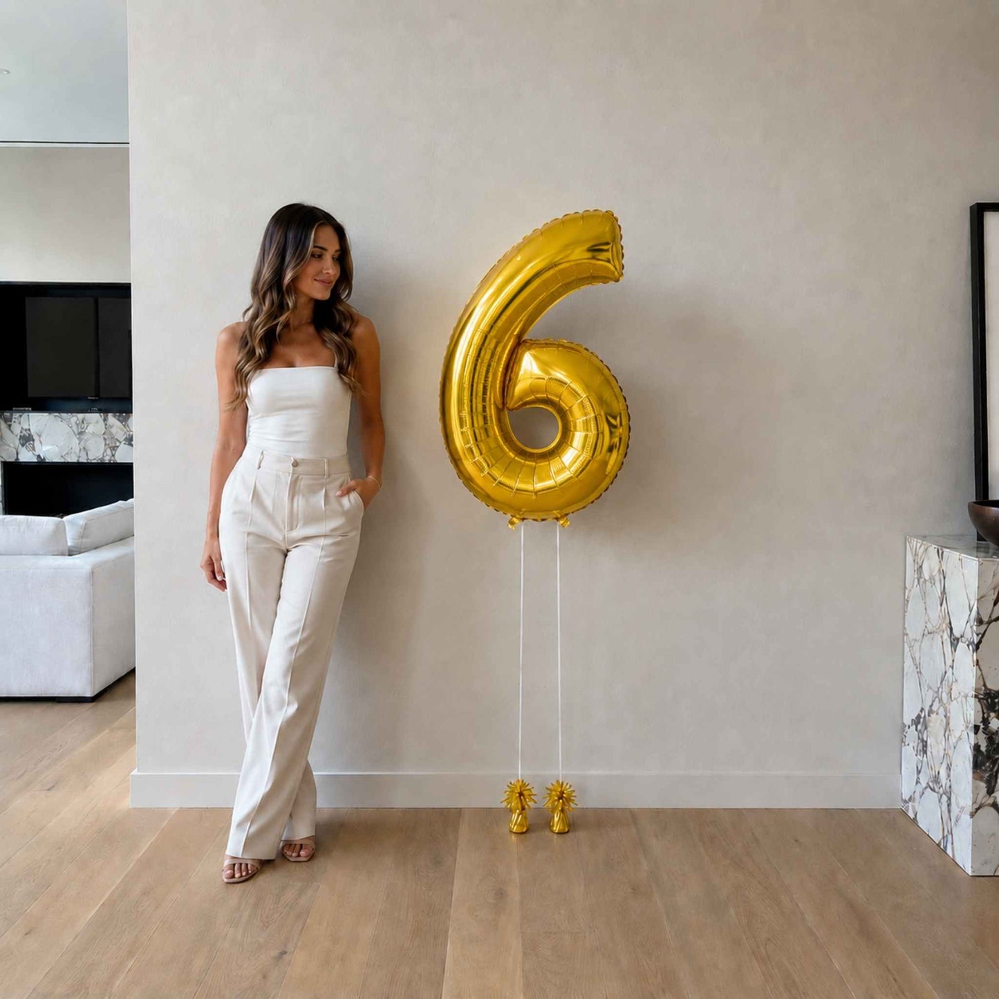 Woman in a white outfit standing next to a large gold number six balloon in a modern living room.