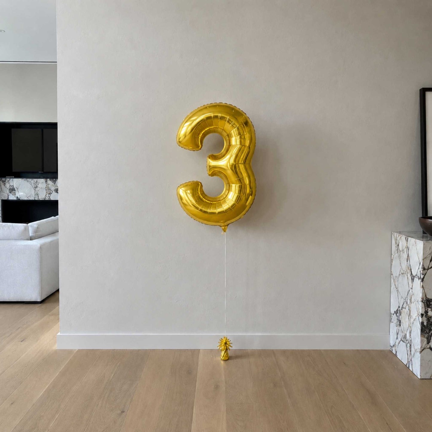 Gold number 3 balloon against a plain wall in a room with a couch and fireplace.