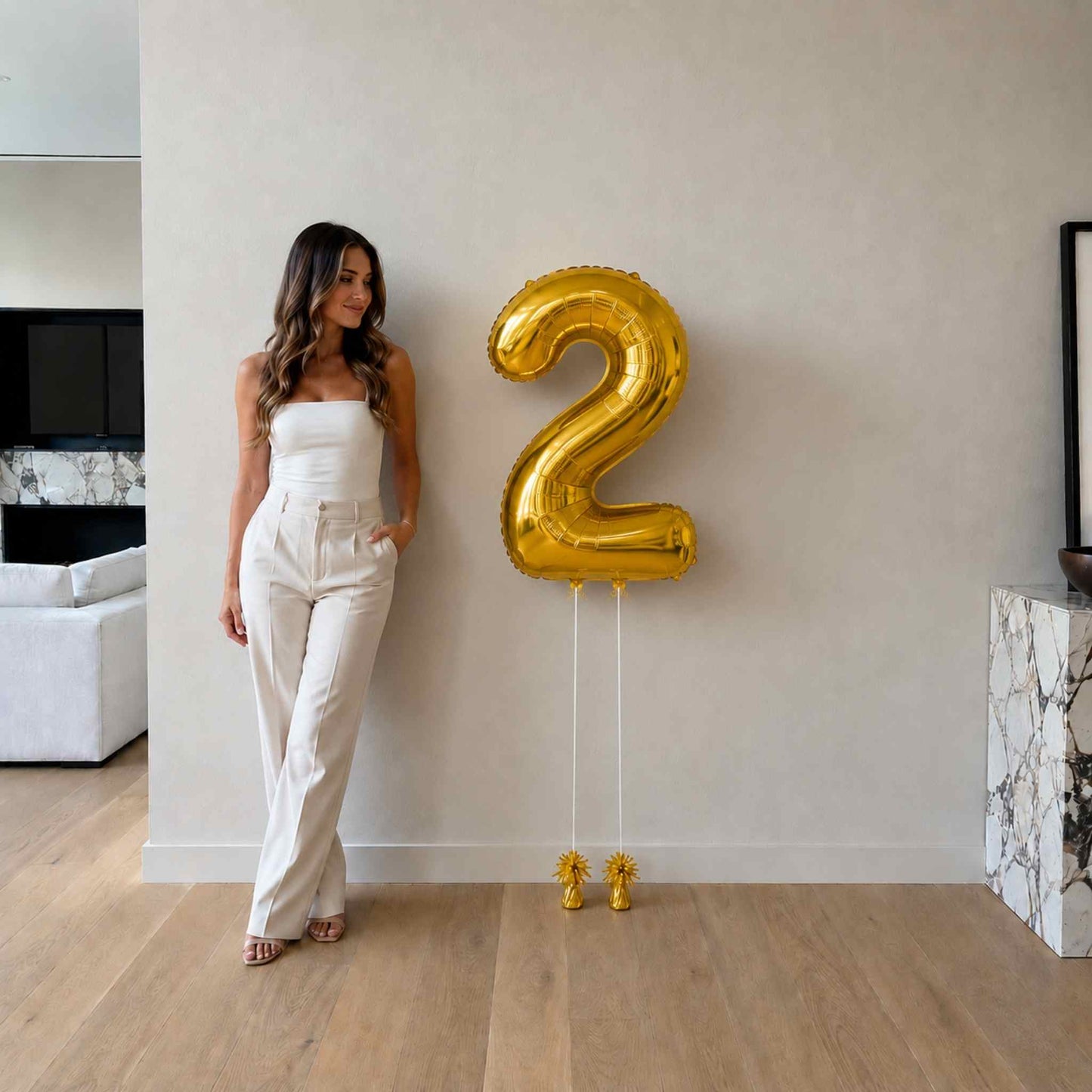 Woman in a white outfit standing next to a large gold number 2 balloon in a modern living room.
