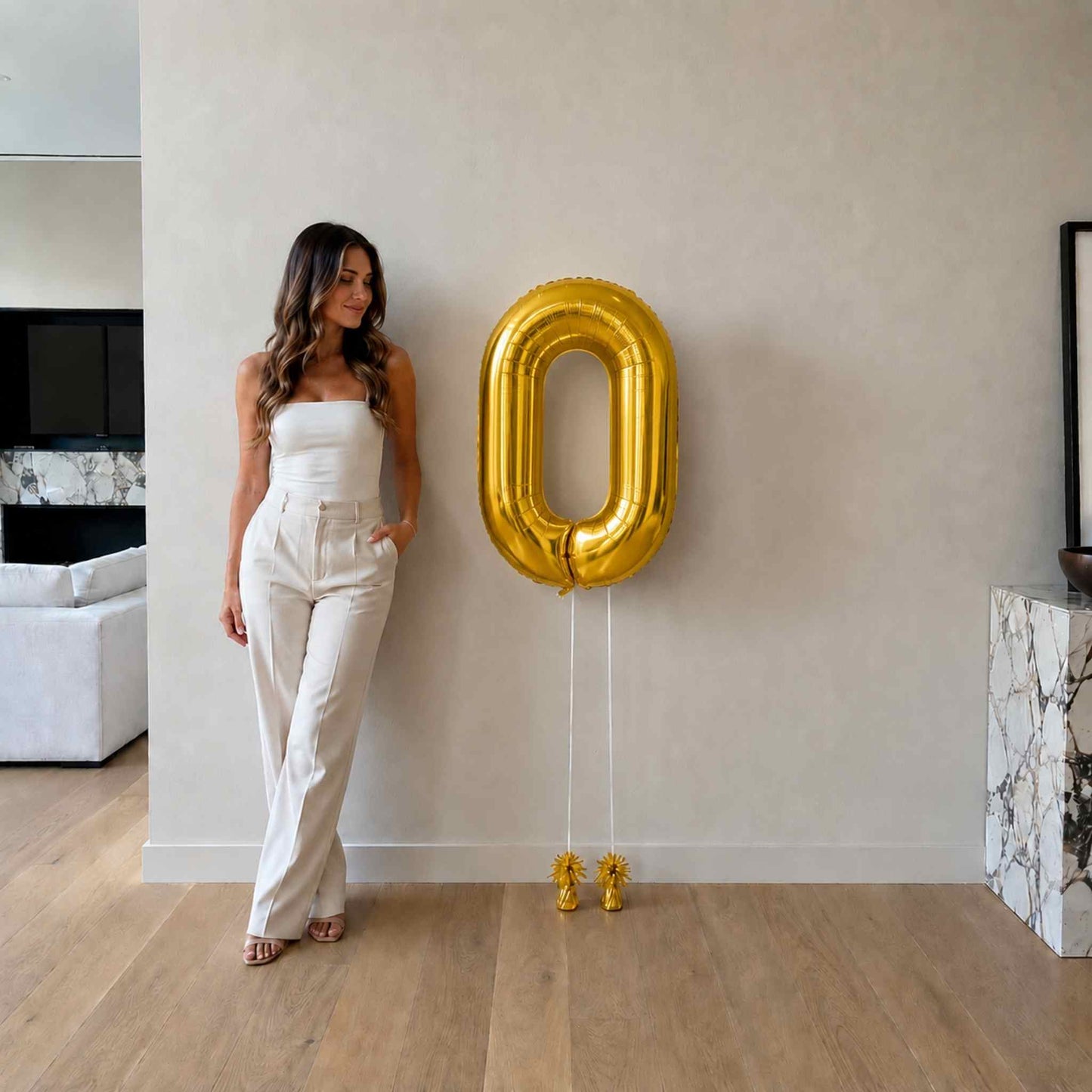 Woman standing next to a large gold number '0' balloon in a modern living room.