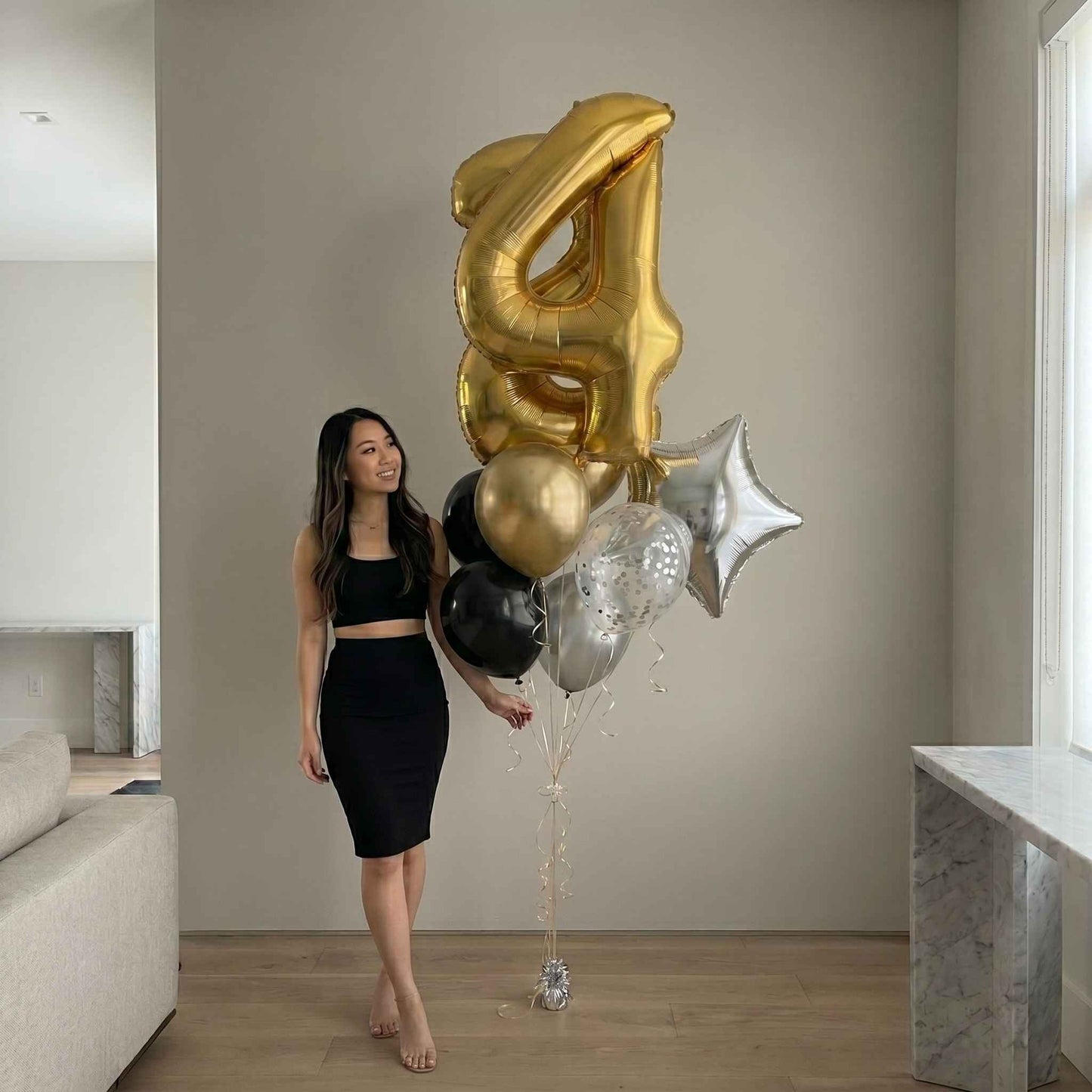 Woman holding a bouquet of gold, black, and silver balloons shaped like numbers in a modern living room.