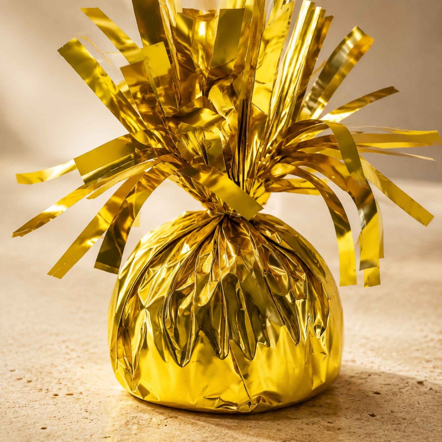 A shiny gold metallic foil-wrapped ornament or decorative bag with a tinsel-like fringe, photographed on a warm beige surface against a softly lit neutral background.