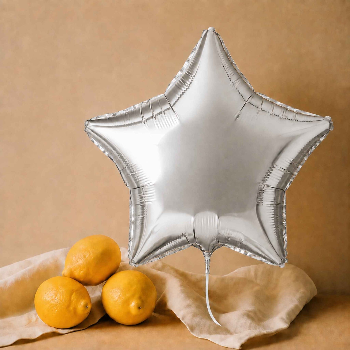A large metallic silver star-shaped foil balloon stands upright on a beige fabric surface, with three yellow lemons beside it against a warm tan background.