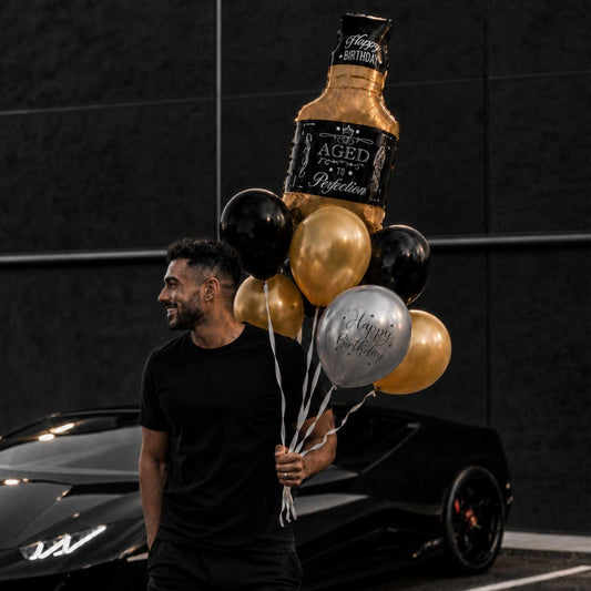A man in a black t-shirt walks past a black sports car while holding a cluster of balloons, including a large gold and black bottle-shaped balloon reading 'Aged 40 Rejection Happy Birthday' and several black and silver round balloons.