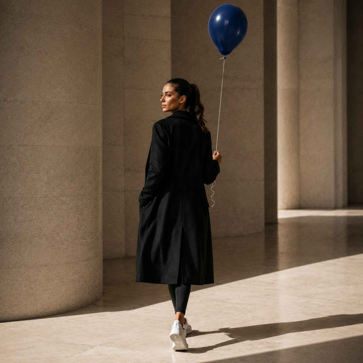 A woman in a long black coat, black leggings, and white sneakers walks away in a sunlit architectural space with large beige columns, holding a blue balloon.