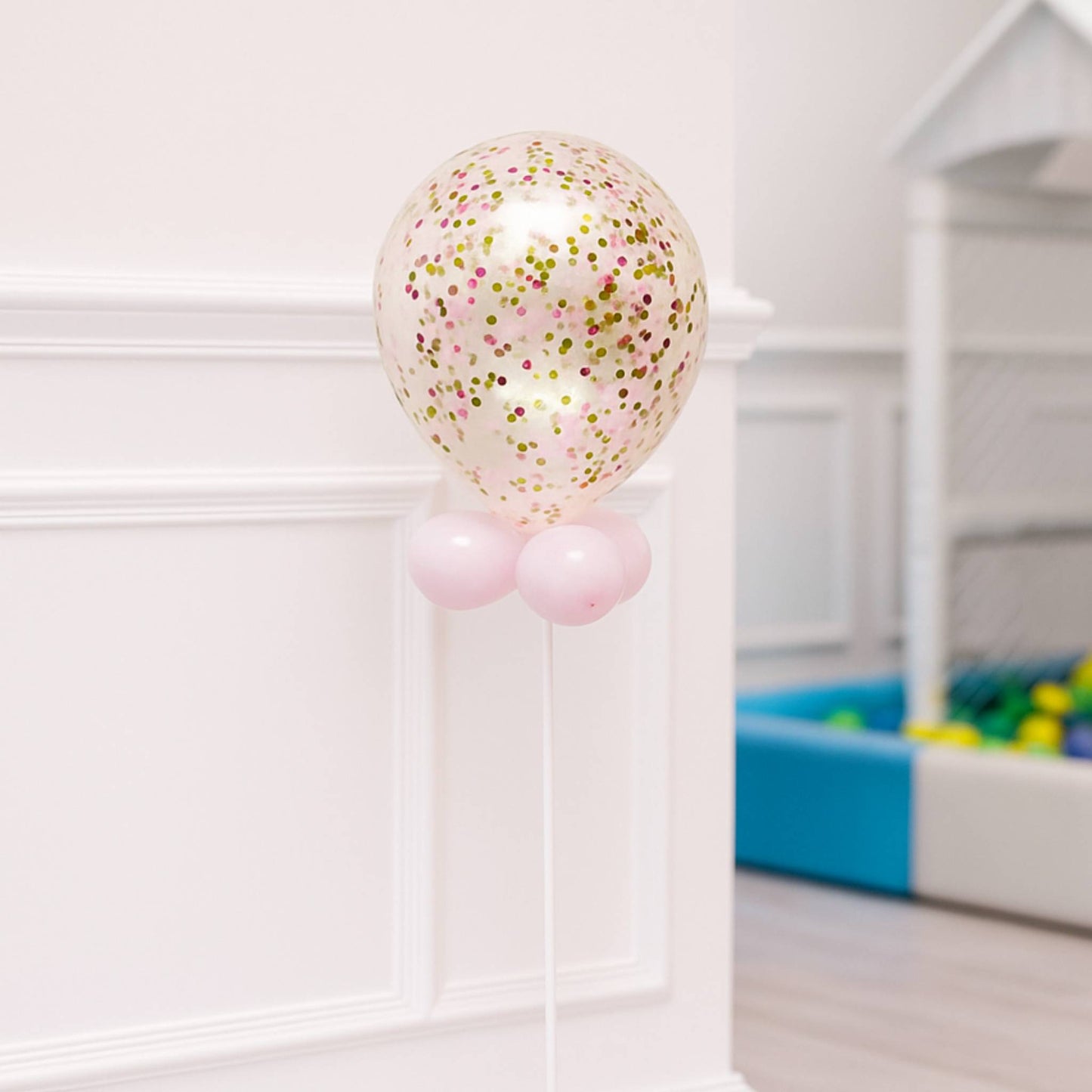 Balloon with confetti and pink base in a room with a white wall and colorful toy box.