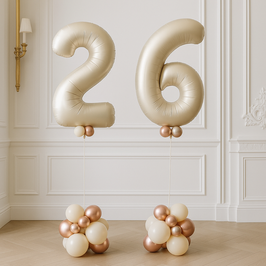 Jumbo gold helium balloons shaped as the number 26 with clusters of smaller gold and white balloons at the base, displayed in a room with white panel walls.