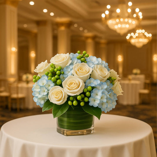 Floral arrangement with blue hydrangeas, white roses, and green berries on a table in a banquet hall.
