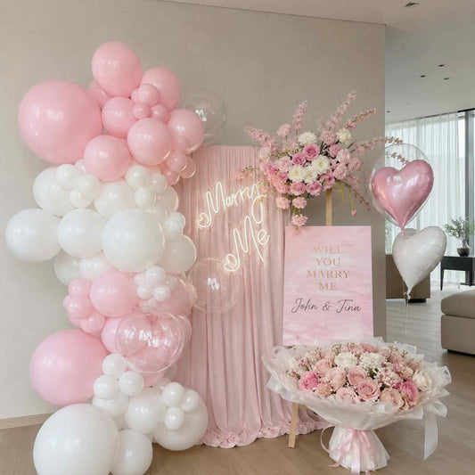 Decorative setup with pink and white balloons, flowers, and a 'Will You Marry Me' sign.