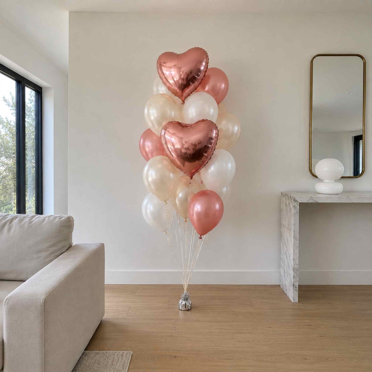 A cluster of metallic heart-shaped and round balloons in rose gold, blush pink, cream, and white, tied to a small stand in a bright living room with a sofa and console table.