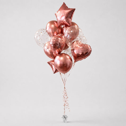 A cluster of metallic rose gold foil balloons in star, heart, and round shapes, mixed with clear balloons containing gold confetti, tied together with a decorative string and a small silver star finial, on a light gray background.