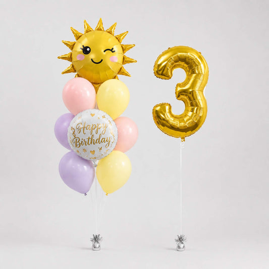 A birthday balloon bouquet and number balloon set on a light gray background. The bouquet includes a gold foil sun balloon with a smiling face, pastel pink, yellow, and lavender balloons, and a round white 'Happy Birthday' balloon.