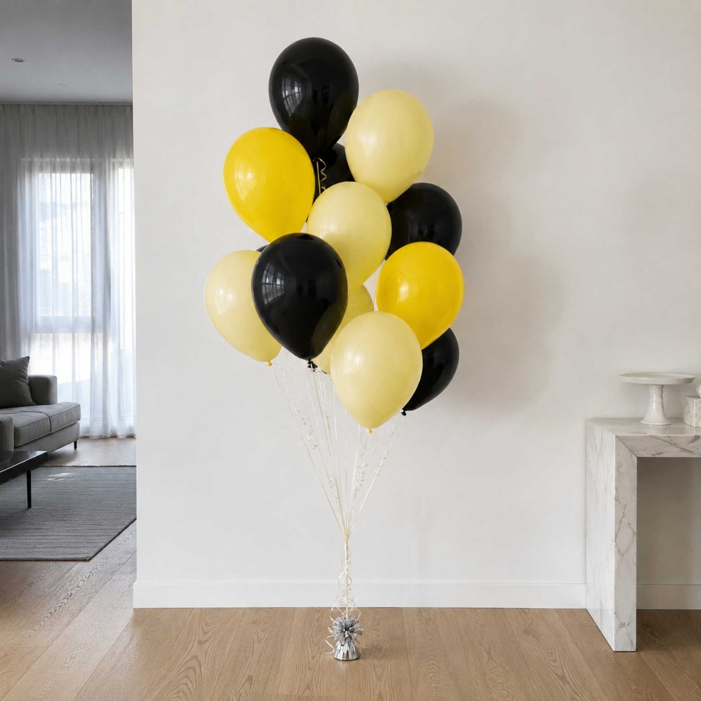 A cluster of latex balloons in black and pale yellow, tied together with clear ribbons and a small silver base, standing on a wooden floor in a bright living room.
