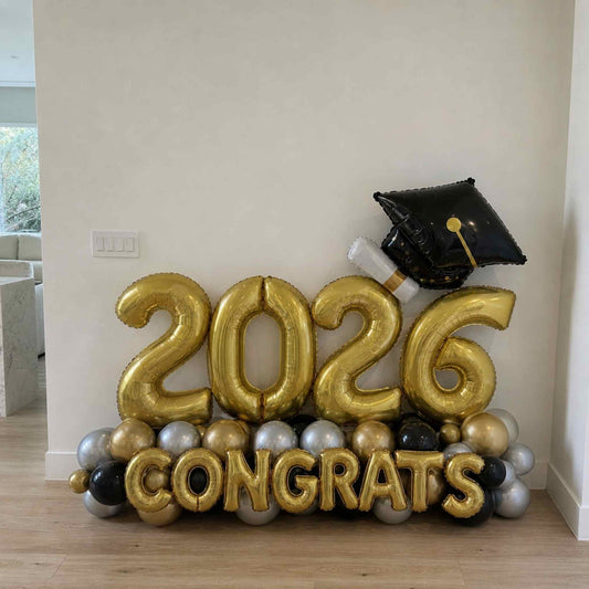 Gold '2026 Congrats' balloons with a graduation cap balloon on a wooden floor.
