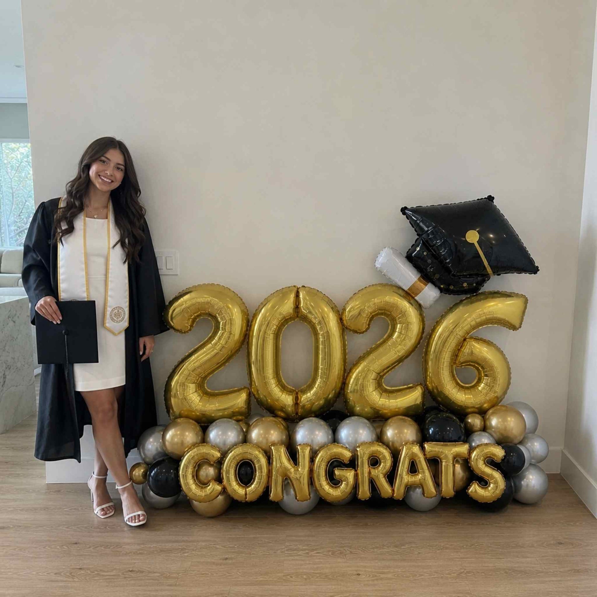 Graduate in cap and gown standing next to '2026 Congrats' balloon display.