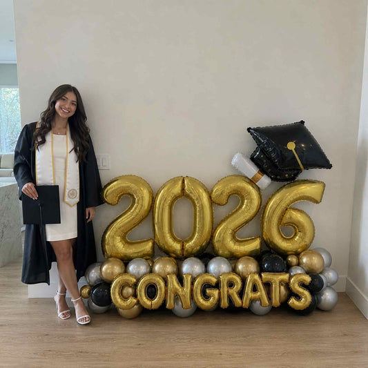 Graduate in cap and gown standing next to '2026 Congrats' balloon display.