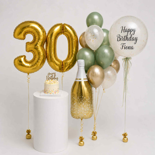 30th birthday party decoration set featuring gold foil balloon numbers '30' on a white pedestal, a champagne bottle with gold glitter, a small birthday cake topper, and a cluster of green, gold, and white balloons including a clear balloon that says 'Happy Birthday Fiona'.