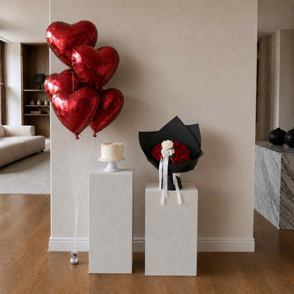 Red heart-shaped balloons, a small cake, and a bouquet on white stands against a beige wall.
