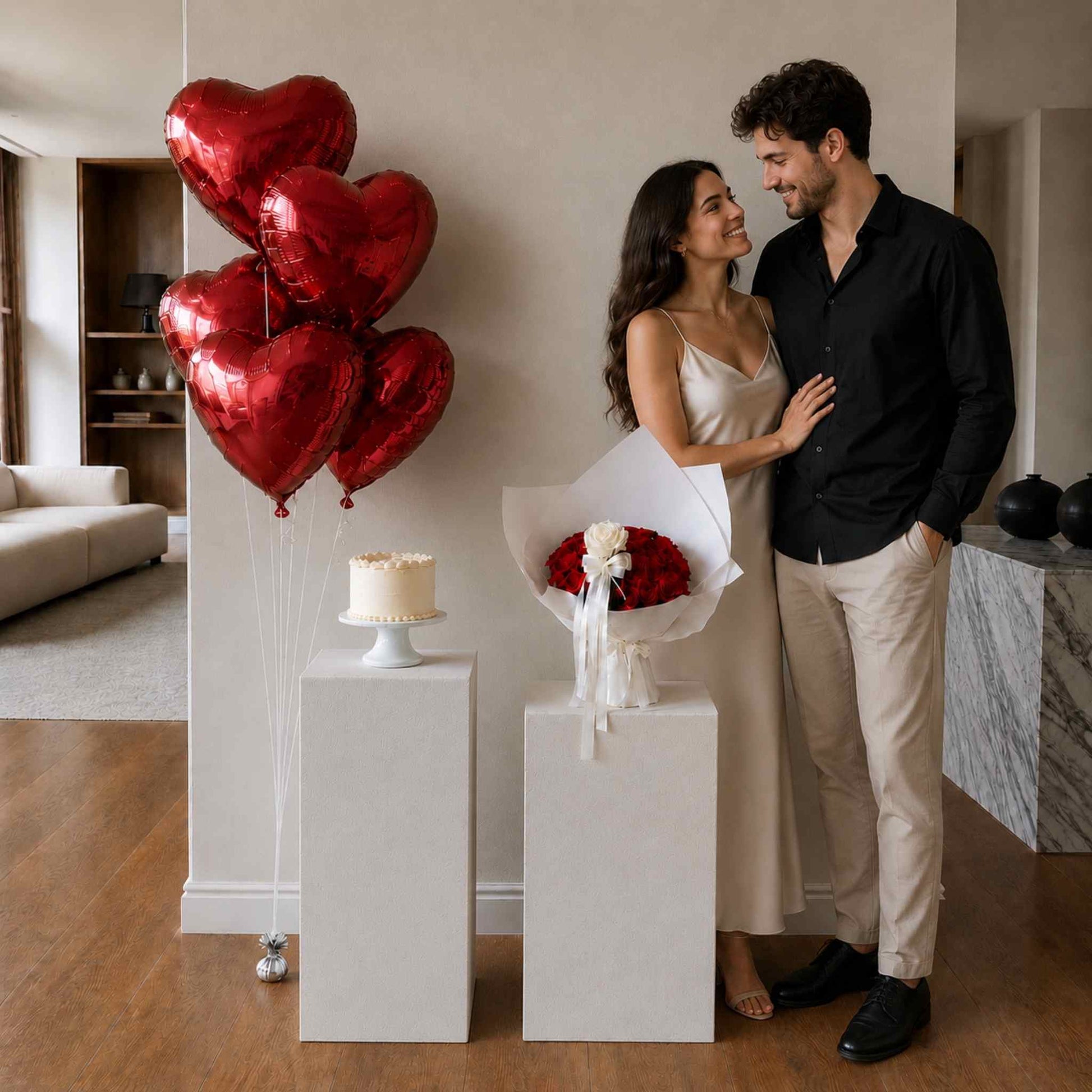 Couple standing together with heart-shaped balloons, a cake, and flowers in a room.