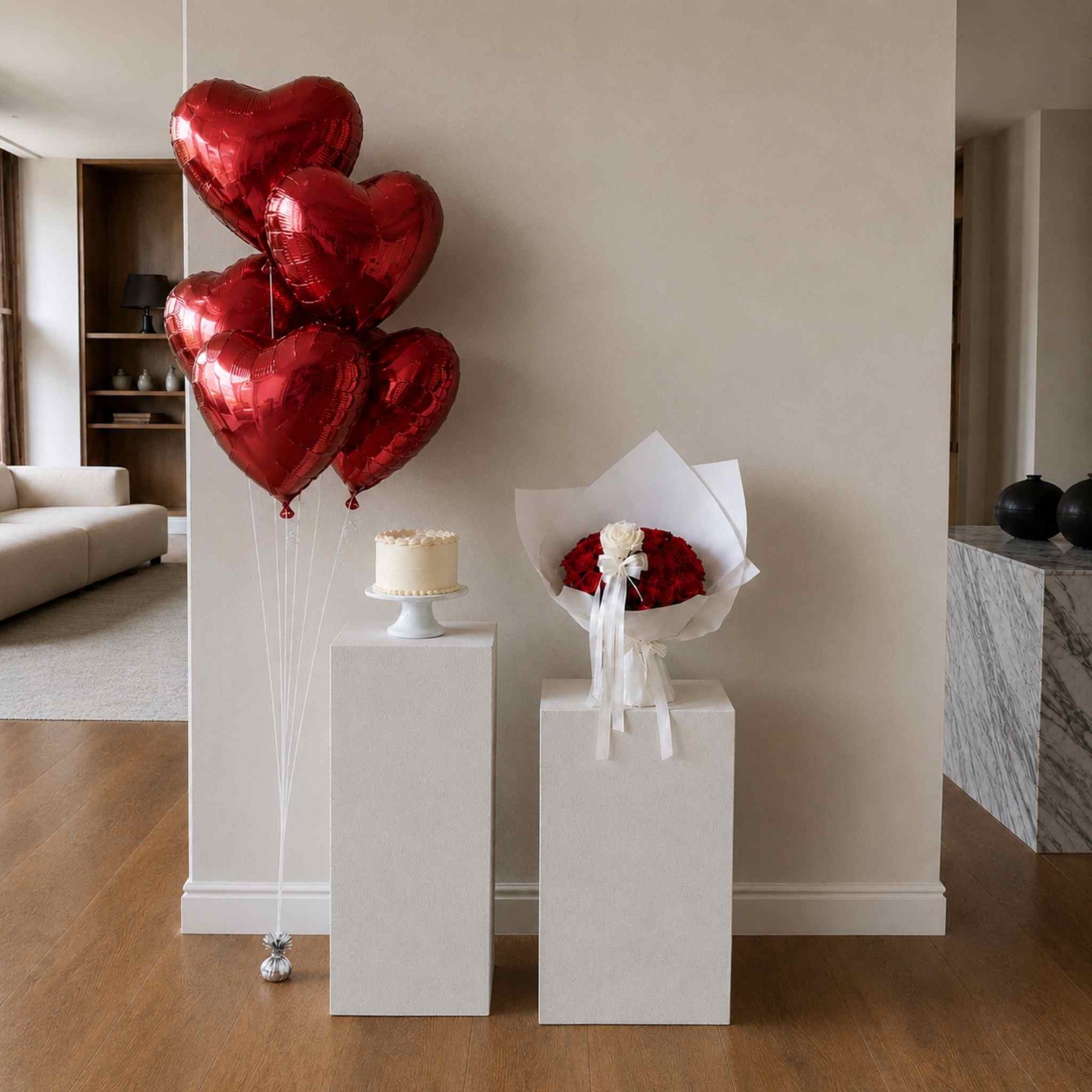 Red heart-shaped balloons and a bouquet on white stands in a room with a couch and shelves.