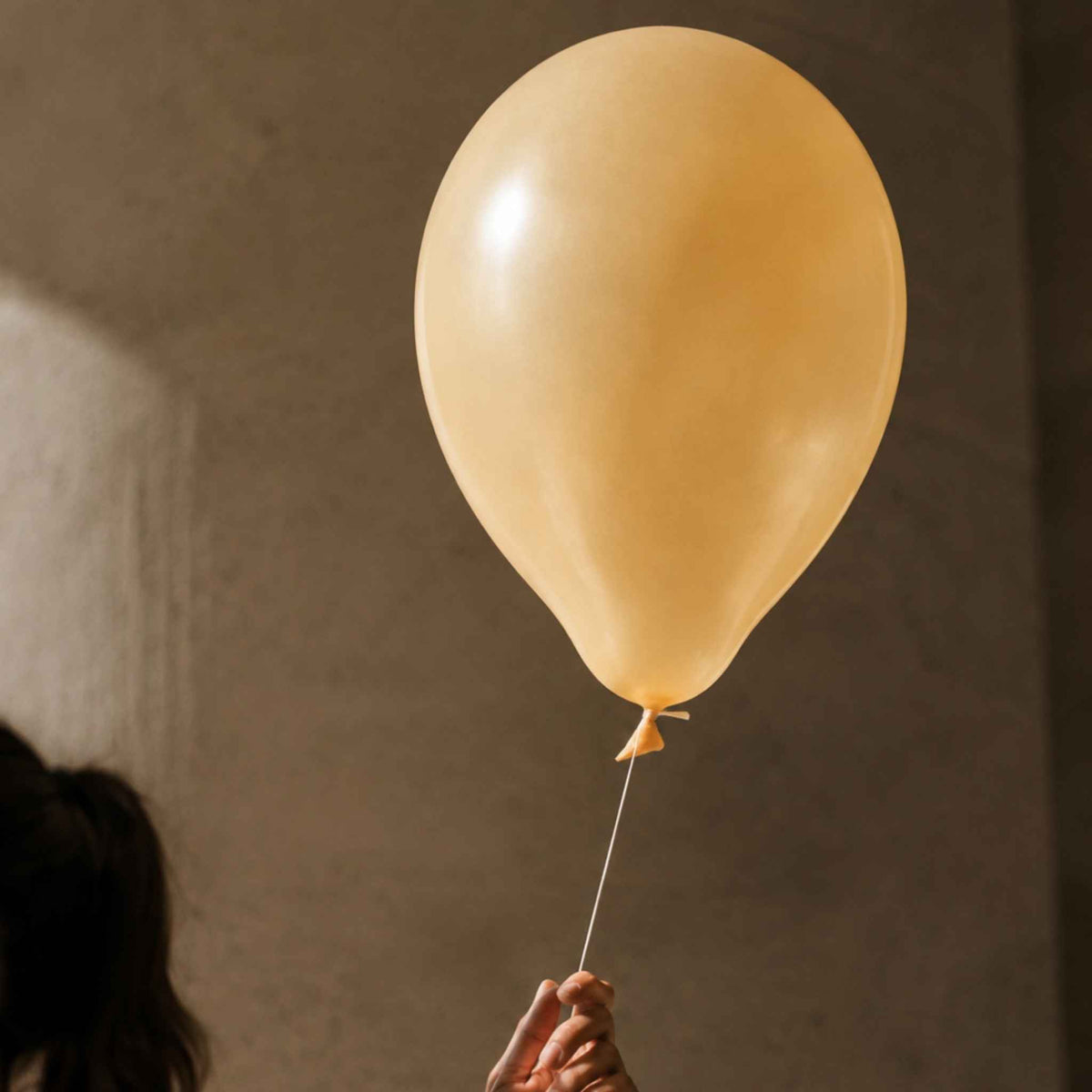 A warm cream-colored latex balloon held by a hand against a textured, warm brown wall.