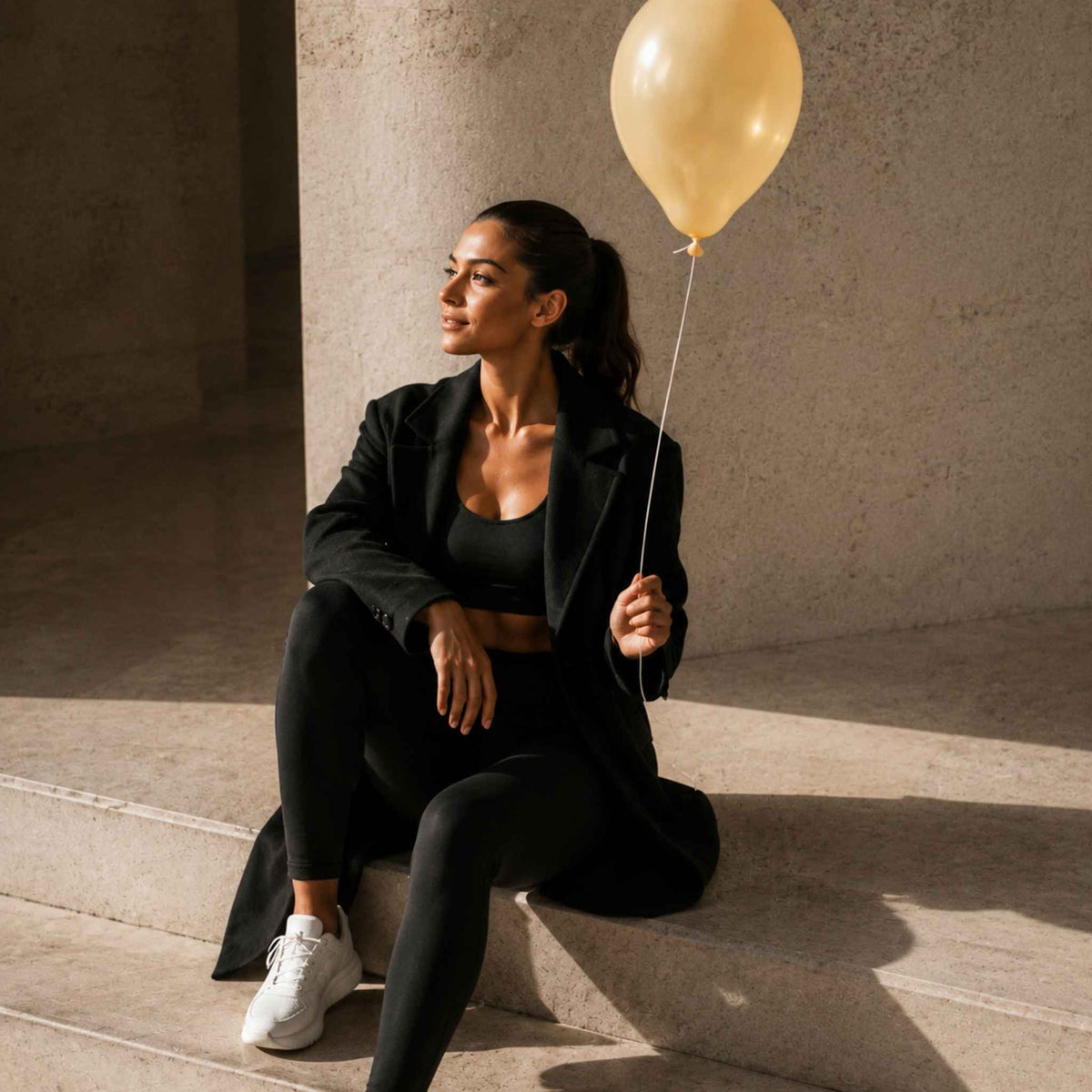 A woman in a black blazer, sports bra, and leggings sits on concrete steps holding a gold balloon, with strong sunlight casting shadows.
