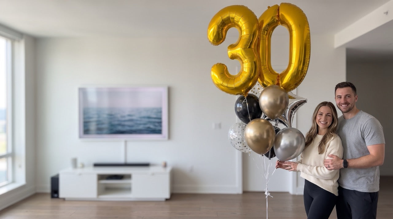 A couple stands in a bright modern living room holding a cluster of balloons, including large gold foil balloons shaped like the numbers 30 and several metallic gold, silver, and black balloons.