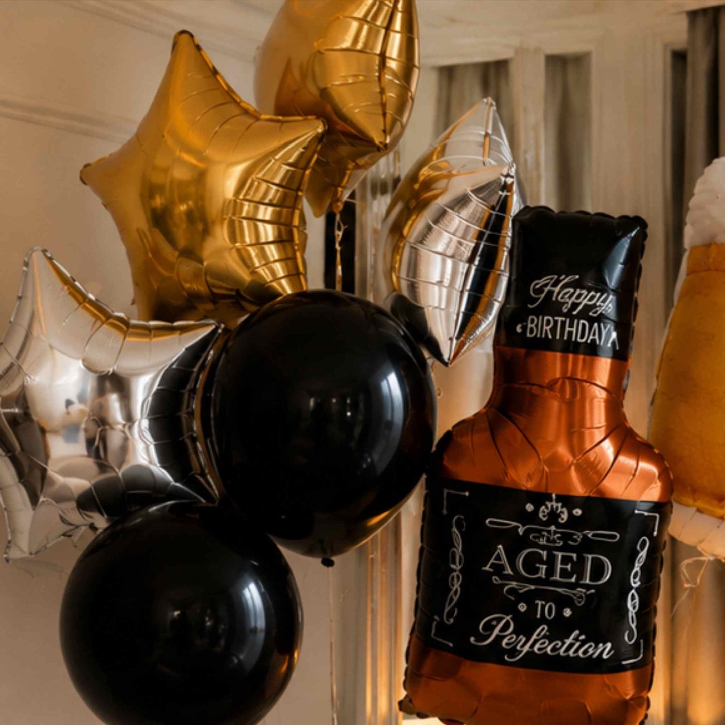 A cluster of metallic and matte party balloons, including gold star balloons, silver foil balloons, two black round balloons, and a bottle-shaped balloon reading 'Happy Birthday Aged To Perfection' in white script.