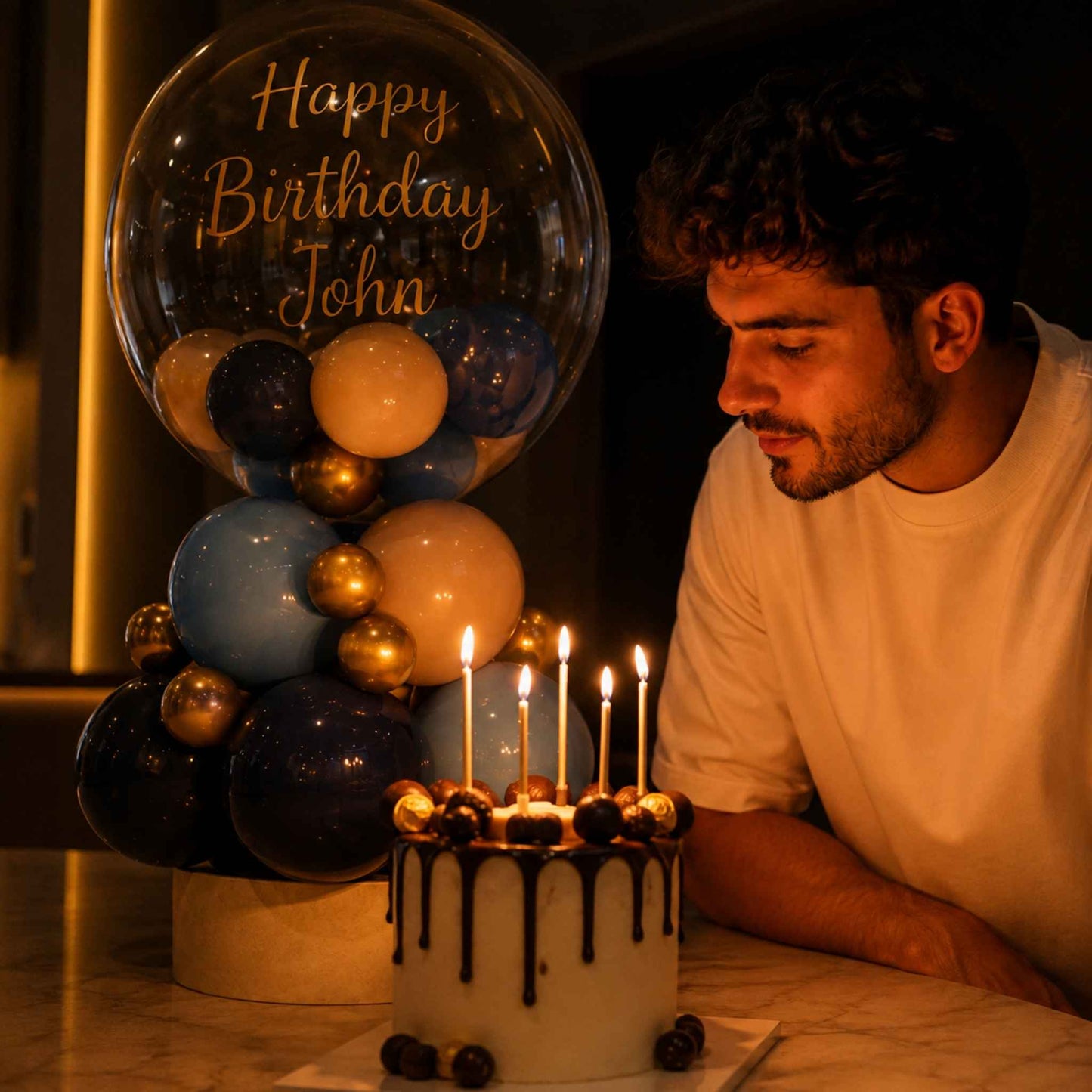 A man in a white t-shirt leans in to blow out candles on a birthday cake next to a large clear balloon that reads 'Happy Birthday John' with navy, light blue, and gold balloons.