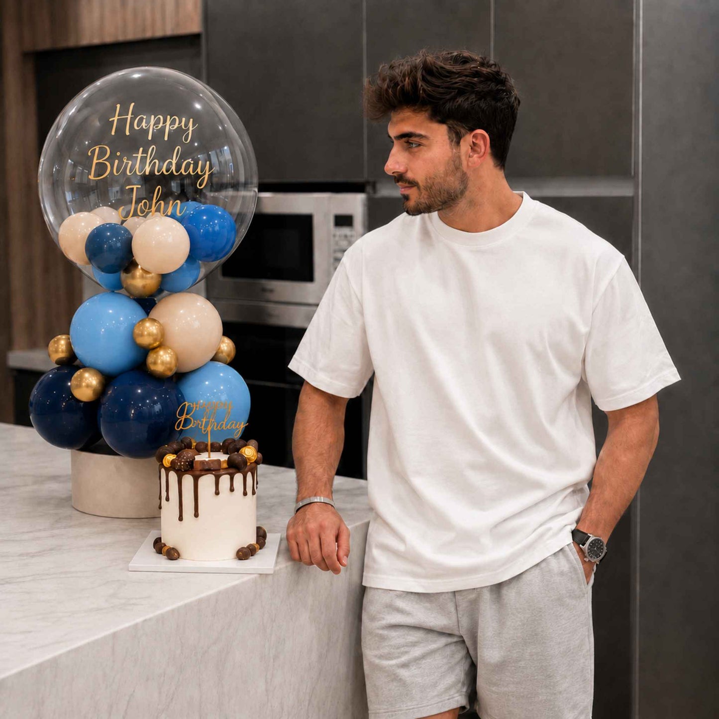 A man in a white t-shirt and grey shorts stands in a kitchen next to a decorated birthday cake and a balloon arrangement with 'Happy Birthday John' written on a clear balloon.