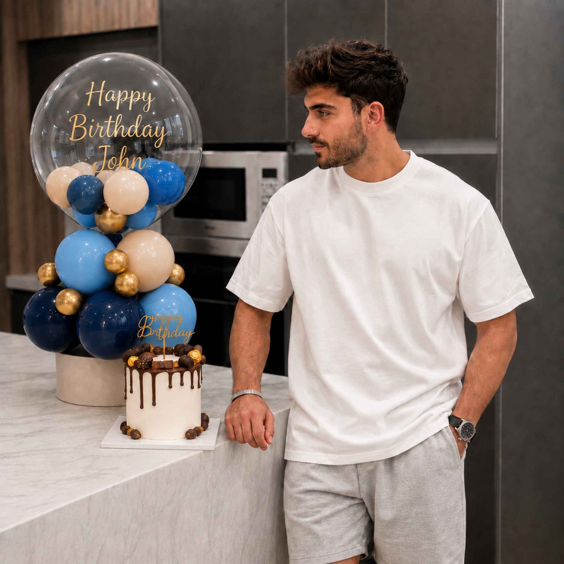 A man in a white t-shirt and grey shorts stands in a kitchen next to a decorated birthday cake and a balloon arrangement with 'Happy Birthday John' written on a clear balloon.