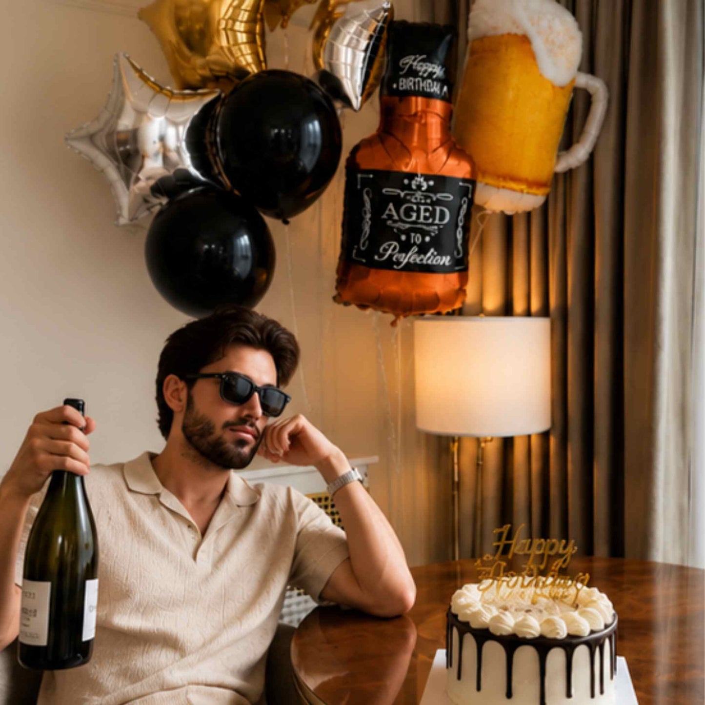 A man in sunglasses sits at a table holding a champagne bottle, with birthday balloons, a cake, and a beer mug balloon decoration in a decorated indoor setting.