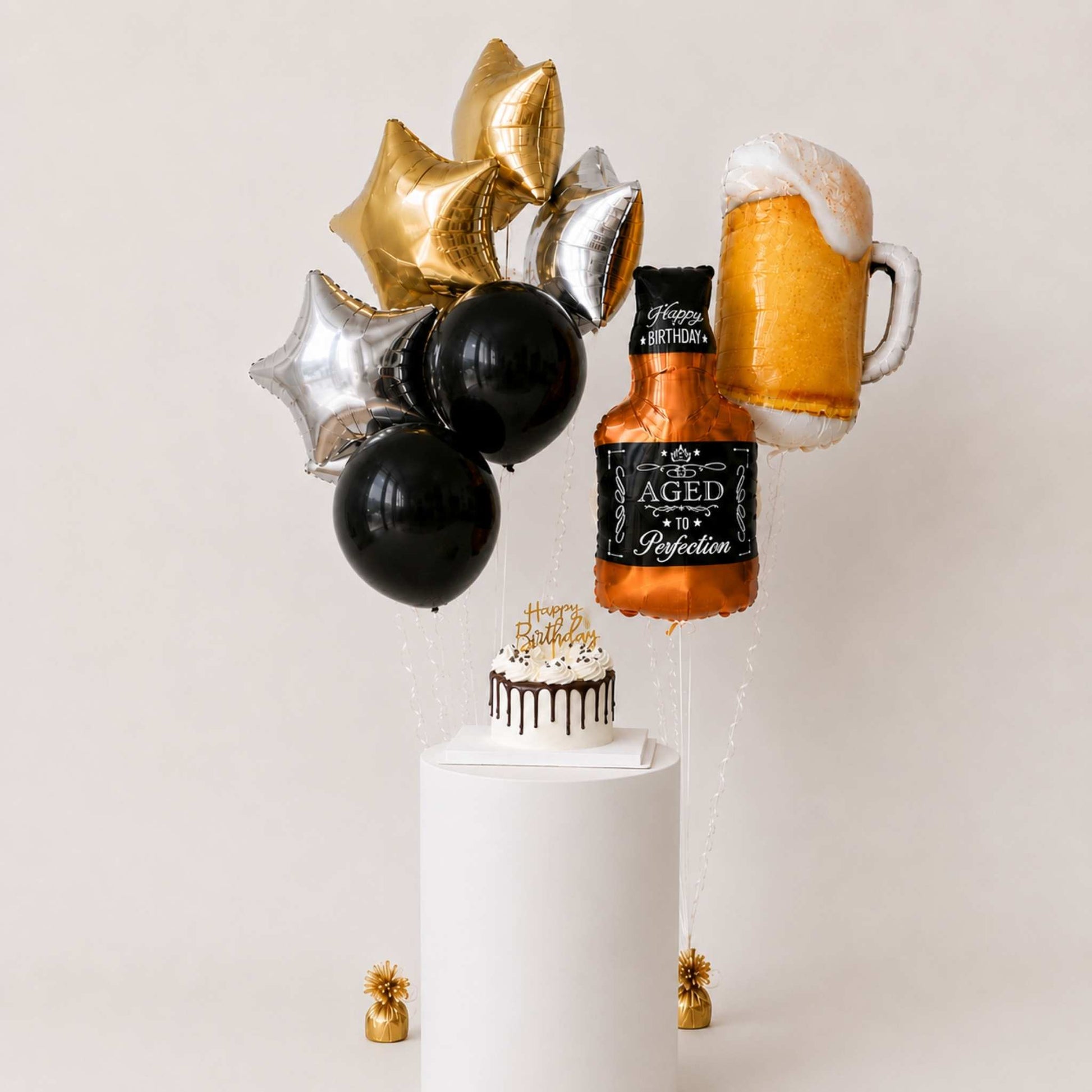 A balloon bouquet centerpiece featuring a whiskey bottle balloon labeled 'Aged 15 Years of Reflection,' a foaming beer mug balloon, black and silver balloons, and a small decorated cake on a white pedestal against a light beige background.
