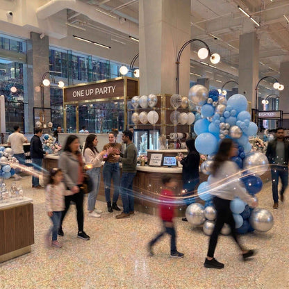 People walking through a modern indoor space with decorative balloons and 'One Up Party' sign.