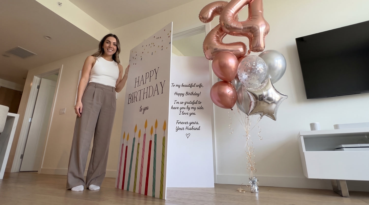 A woman stands in a modern living room next to a large personalized birthday sign that reads 'Happy Birthday to you' with candle graphics, alongside a balloon bouquet with metallic rose gold and silver balloons.