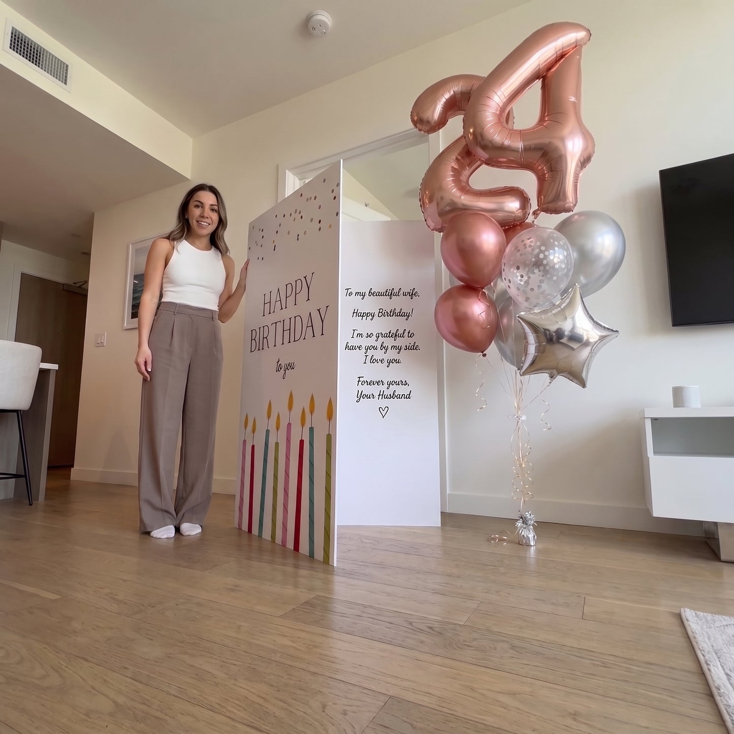 A woman stands in a living room next to a large birthday sign that reads 'Happy Birthday' with candle graphics, alongside a cluster of balloons including a large metallic rose-gold balloon shaped like the number 4 and other metallic balloons.
