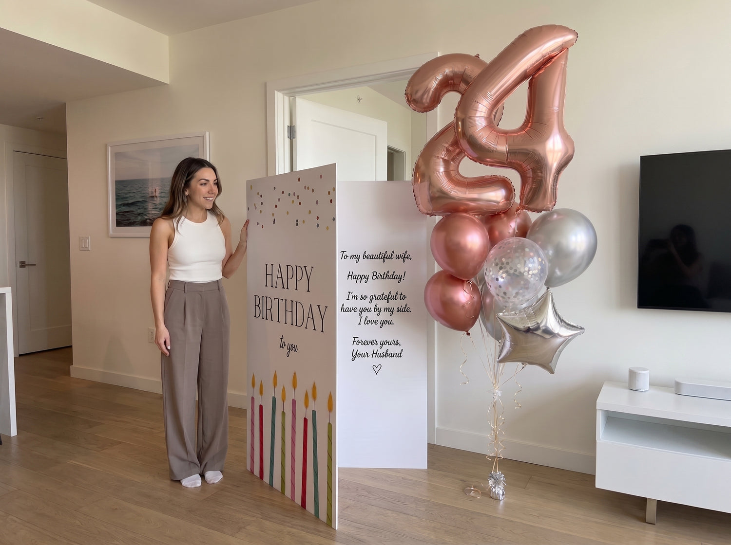 A woman stands beside a birthday balloon decoration setup in a home interior. The setup includes a large metallic rose-gold balloon shaped like the number 4, a cluster of rose-gold and silver balloons, and two tall printed panels with birthday text and candle graphics.