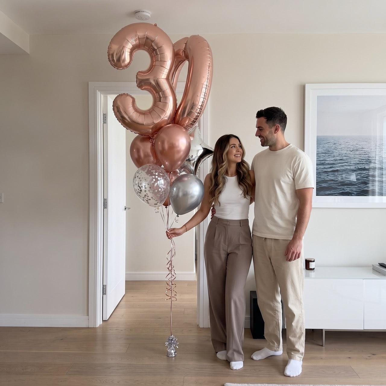 A couple standing in a bright, modern room holding a large balloon bouquet with metallic rose gold number balloons and silver balloons.