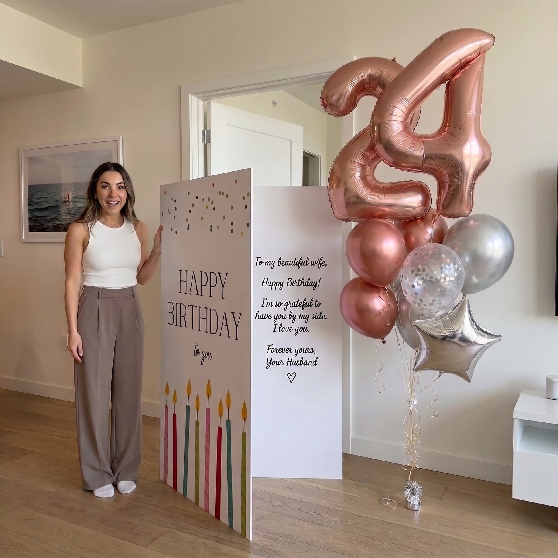 A woman stands indoors next to birthday party decorations, including a large freestanding 'HAPPY BIRTHDAY' sign, a white message card, and a cluster of metallic balloons in rose gold, silver, and clear with a large number 24 balloon.