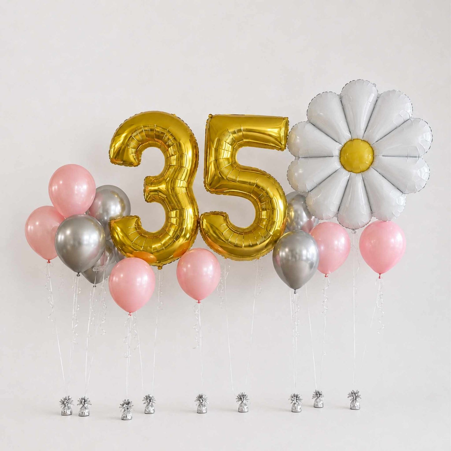 A balloon decoration set for a 35th birthday, featuring metallic gold number 35 balloons, a white daisy balloon with a yellow center, pink and silver balloons, and small silver foil cupcake toppers on a white background.