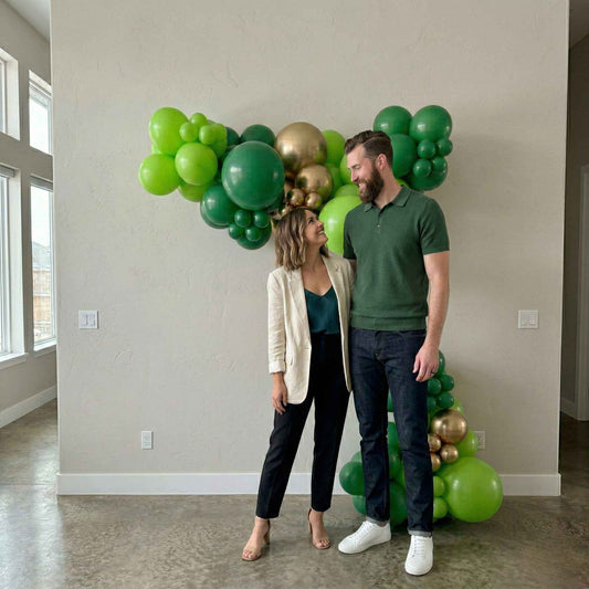 Man and woman standing in a room with a large balloon garland arrangement of green and gold balloons.